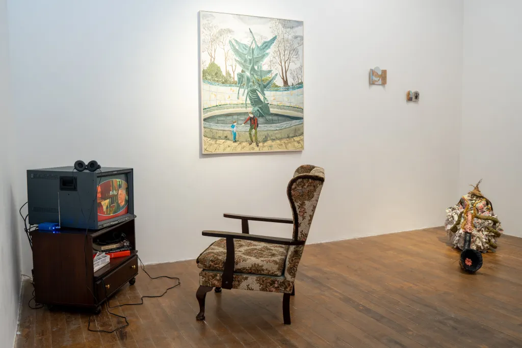 Exhibition documentation of a white wall gallery with wooden floor, an old TV on a Wooden stand containing books underneath sits in the left corner with an old wood and floral print beige armchair facing it, a large painting with two smaller paintings hang on the wall in the middle and a small abstract sculpture of a creature playing an instument sits on the floor to the right.