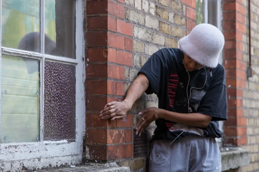 Peformance documenation, a person wearing headphones is standing hunched over in front of a buliding, their two hands reach around holding the red brick framing the window. 