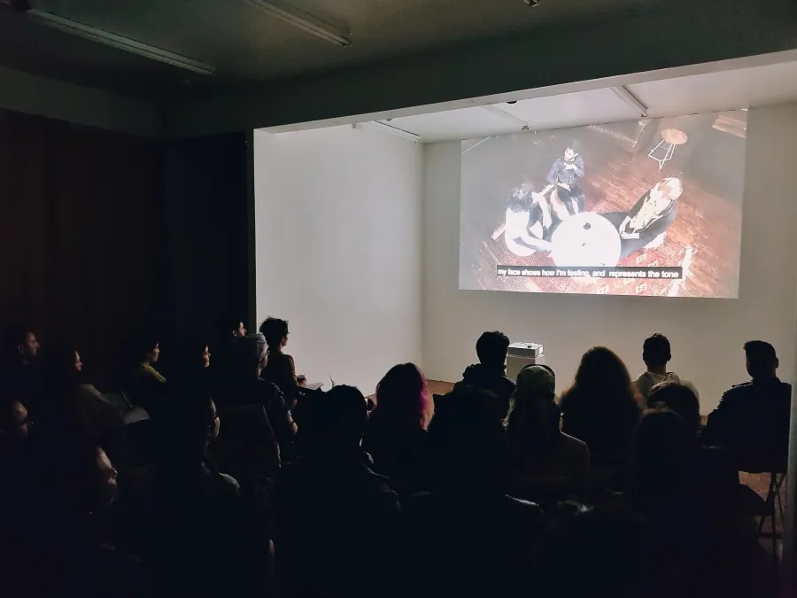 Documentation of a screening, a large group of people sit in a darkened room watching a projected video depicitng an overhead shot of a group of 3 people sitting around a small circle table.