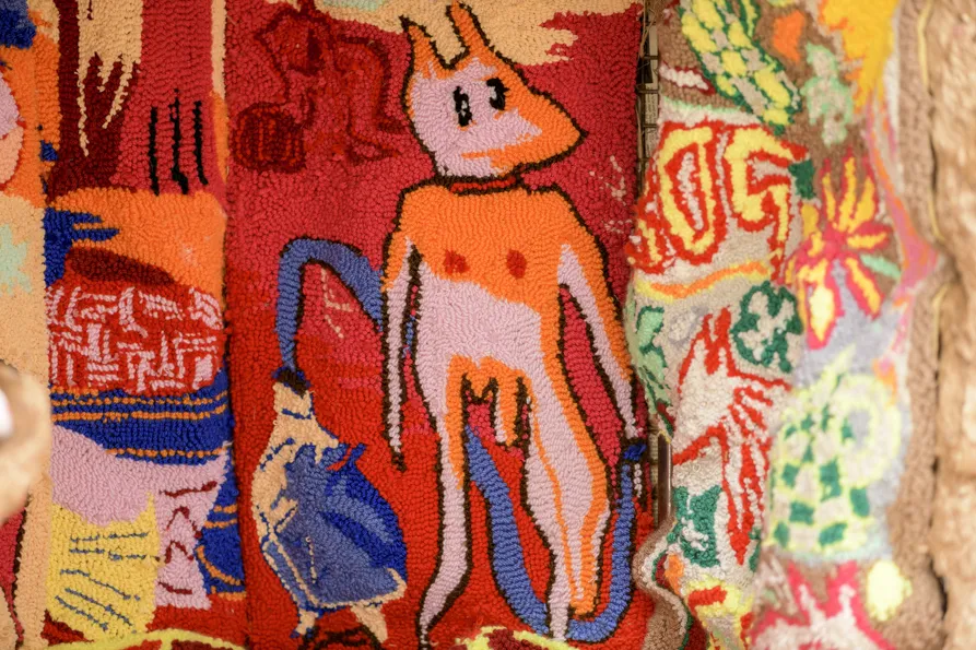 Close up documentation of a carpet piece with an abstract image of a dog-like person in pink and orange surrounded by various shapes in multicolured red, pink, orange and blue background