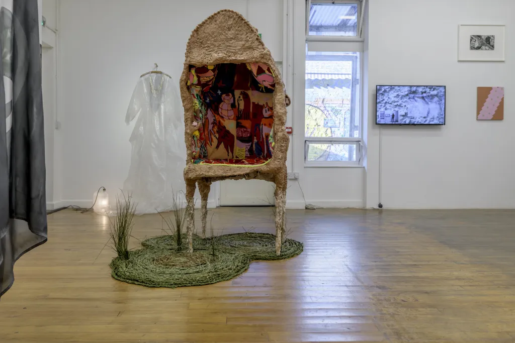 Exhibiton documentation in a white wall gallery with a wooden floor, a large sculpure stands on the floor it has four legs and holds a carpet with various images in a cave like opening, underneath is a circular green carpet with tuffs of long grass.