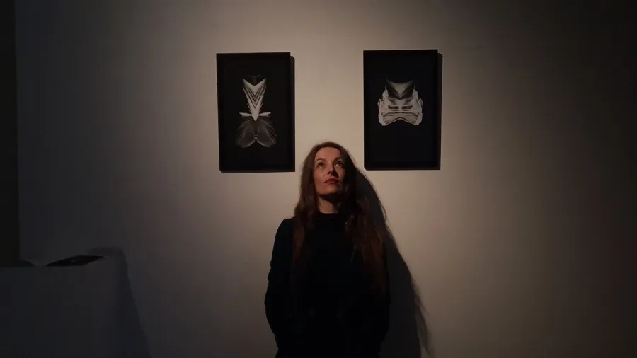 A woman dressed in black stands below two black and white abstract prints hanging on a white wall in a darkened room, she faces the camera and is looking up.