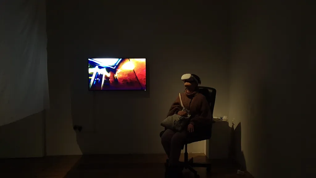 Exhibition documentation of a darkened gallery space with white walls, a person sits on chair looking up and wears a VR headset, a TV displaying a live view of the VR hangs on the wall beside her. 