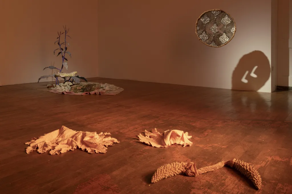 White wall gallery with a wooden floor and dusty orange lighting, installation view with abstract sculptures, suspended circular object and an arrangement of sculptures on the floor ressembling organic and natural shapes.