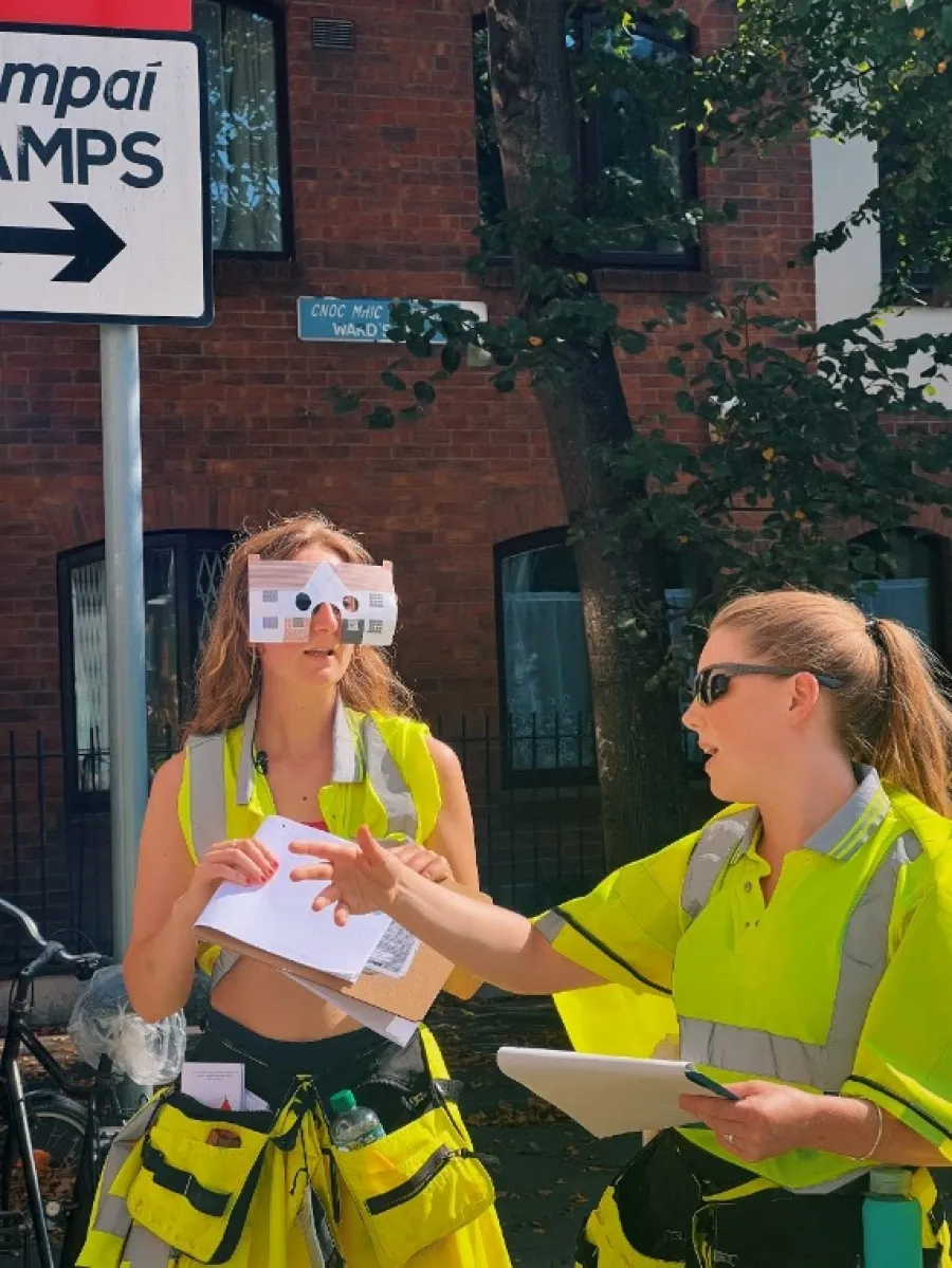 Two people talking, wearing visibitity jackets, holding clipboards. One wears a paper house fashioned as glasses. Brick houses, a tree and a street sign as background.