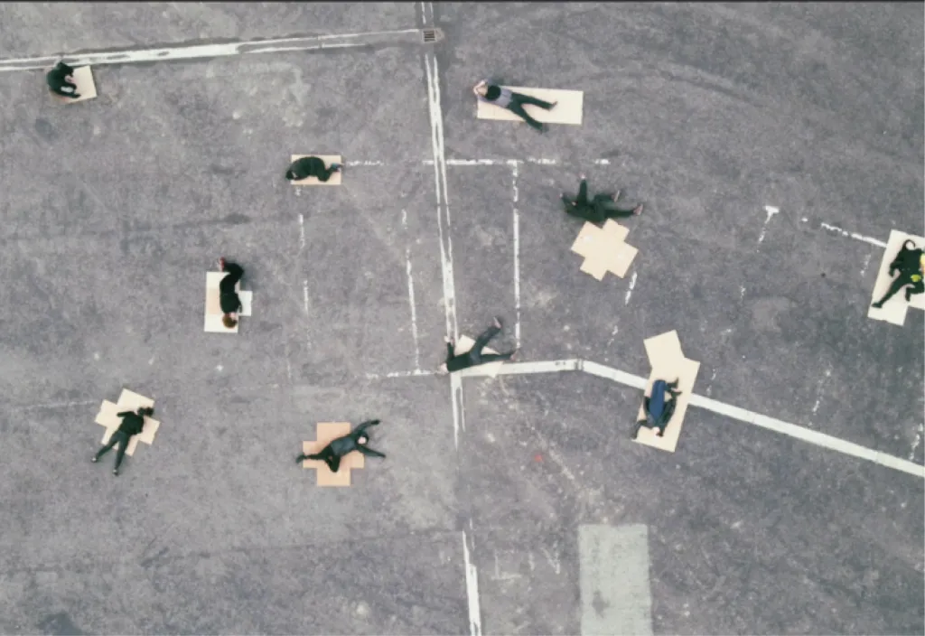 Aerial view of a concrete ground, maybe a old parking lot, where 10 people lay down on carboard in different fashions.