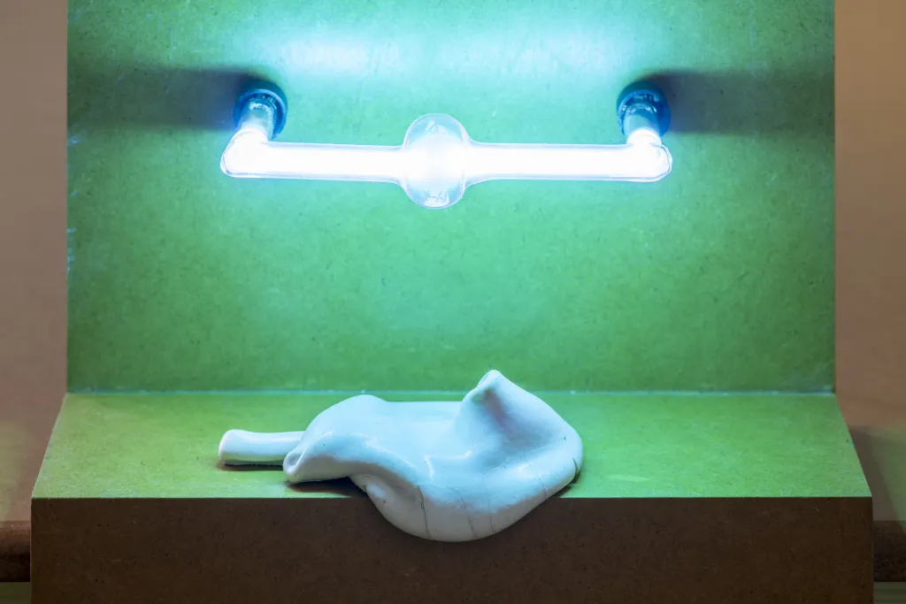 A sculpture of a green shelf-like object, a cermaic white object ressembling a deflated ballon hangs over the edge and a fluorescent white light is attached to the back.