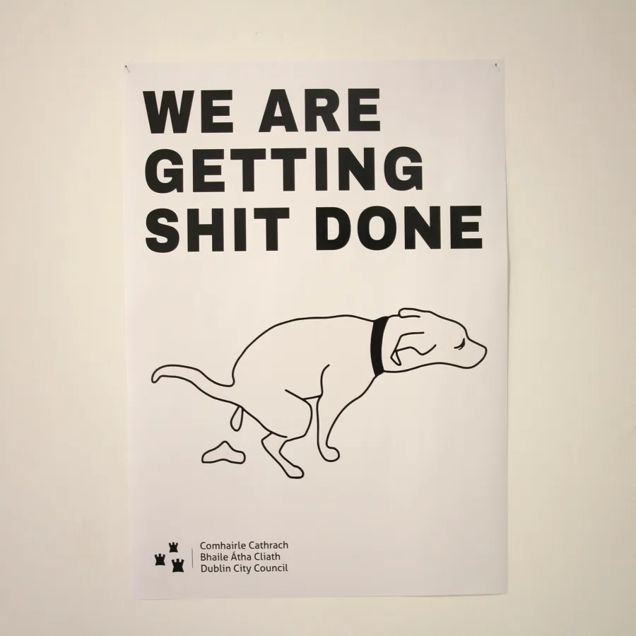 A white A4 poster that reads 'We are getting shit done' in large black font with an image of a line drawing of a dog deficating, with the Dublin City Council logo in the bottom left corner, on a white wall.