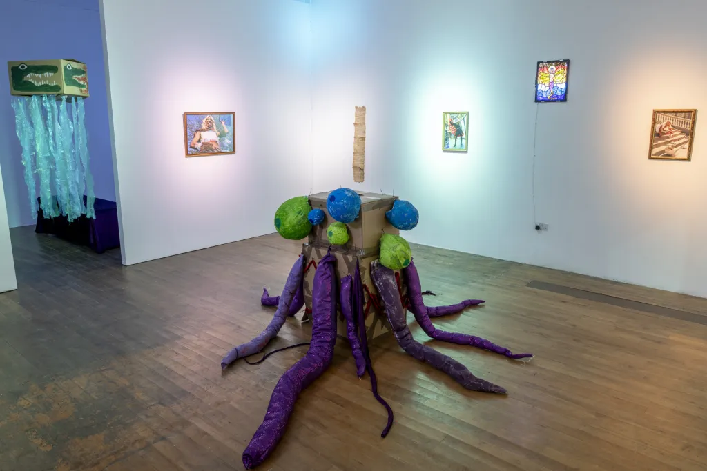 Exhibition documentation of a white wall gallery with wooden floor, a sculpture of a cardboard box with purple tenticles sits in the middle of the room, photographs and a long script hangs on the walls, a box with a crocodile heads painted on and blue tenticles hangs from the ceiling. 